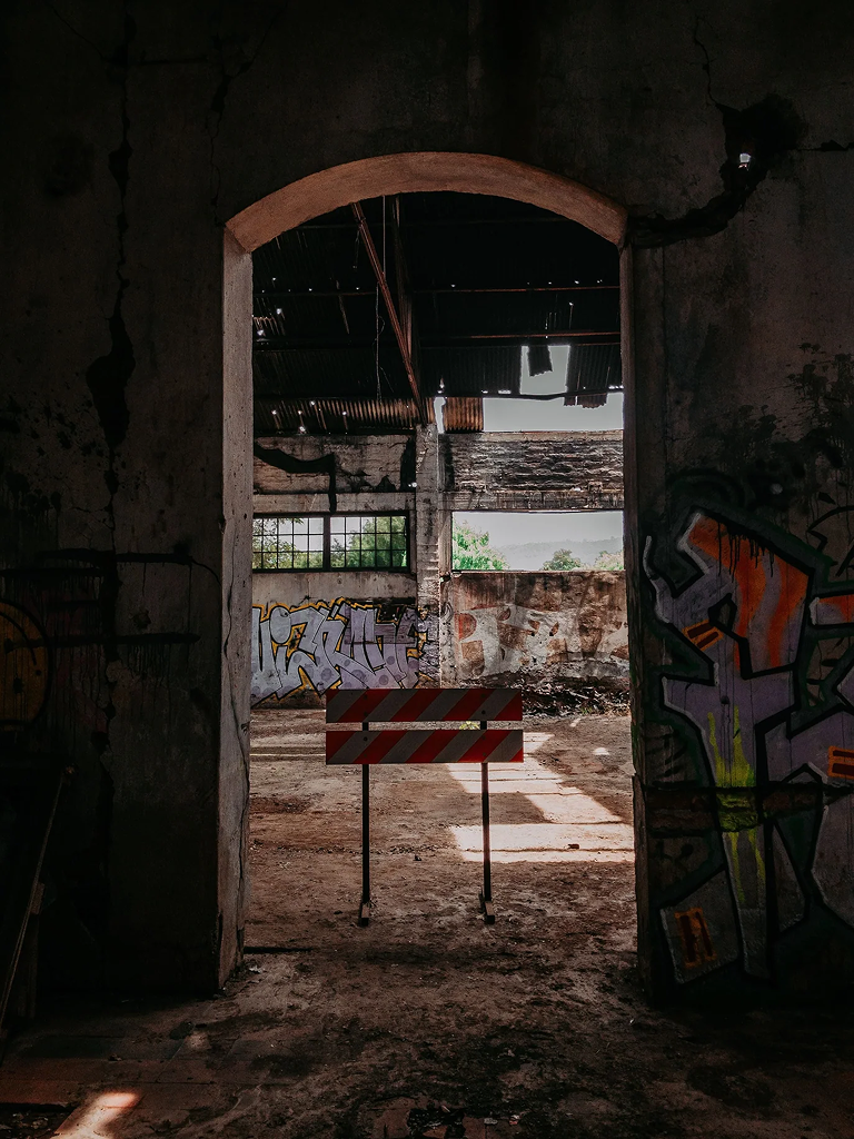 Arched doorway in an abandoned, graffiti-covered building, with a striped barrier and sunlight streaming through a damaged roof.