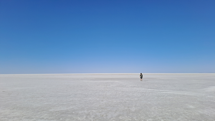 A lone person with a backpack and hat stands small on a vast, flat white salt plain beneath a wide clear blue sky stretching to the horizon.