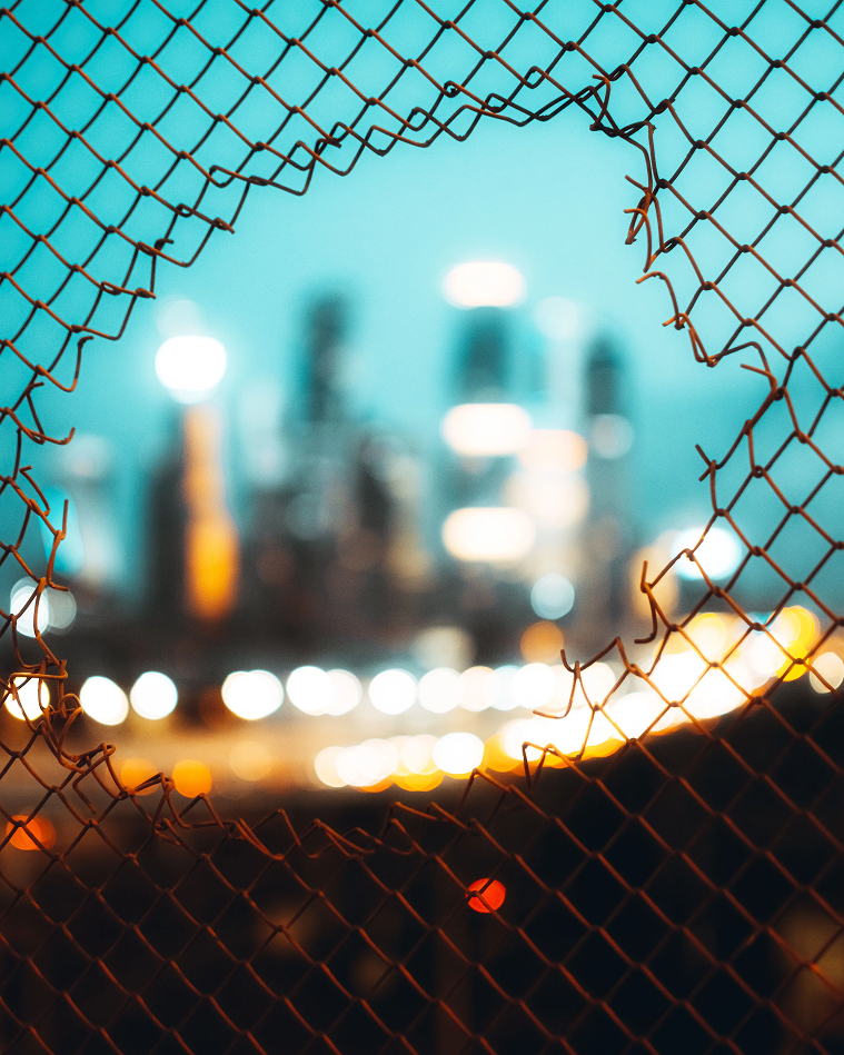 A hole in a chain-link fence frames a blurred city skyline at dusk.