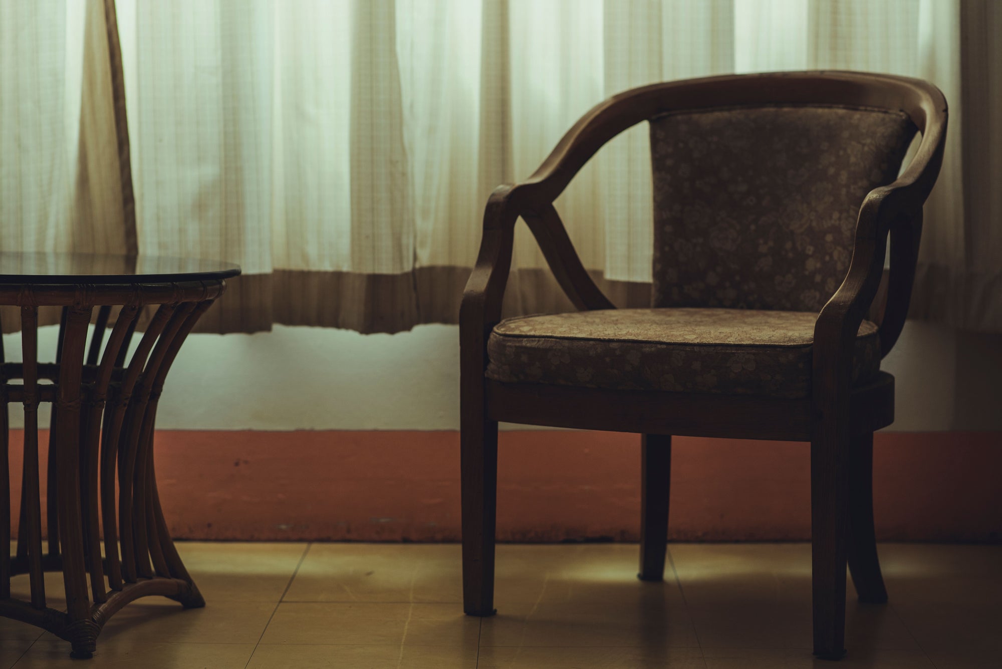 An empty vintage chair beside a small table and softly lit curtains.