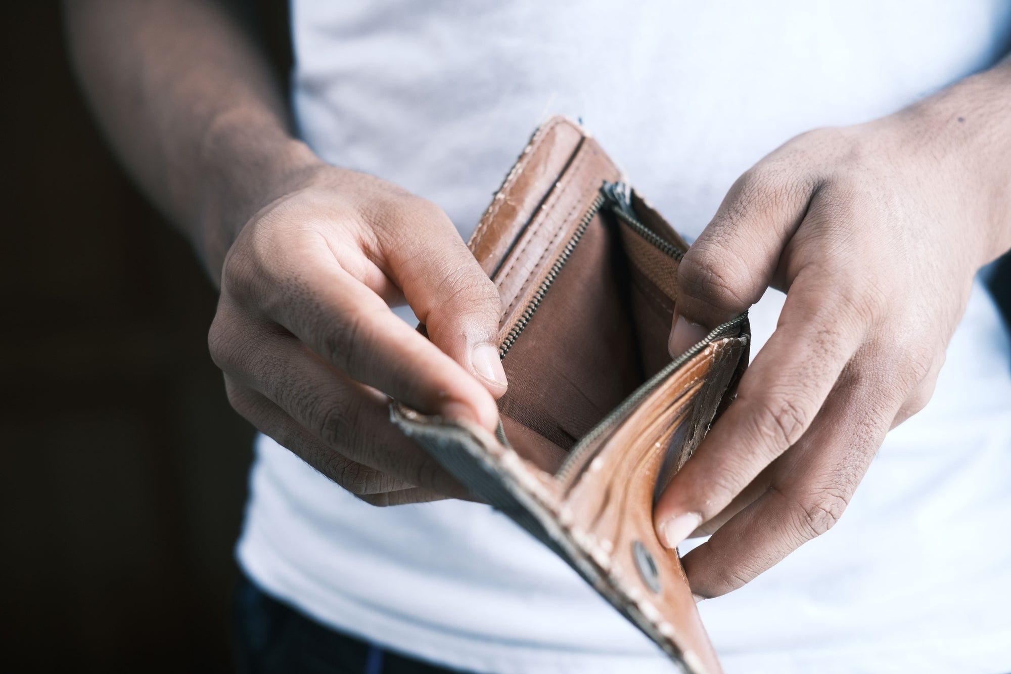 A pair of hands holds open an empty brown leather bifold wallet, pulling the zippered compartment apart to show nothing inside. The person wears a white t-shirt, blurred in the background, with the focus on the worn wallet and their hands.