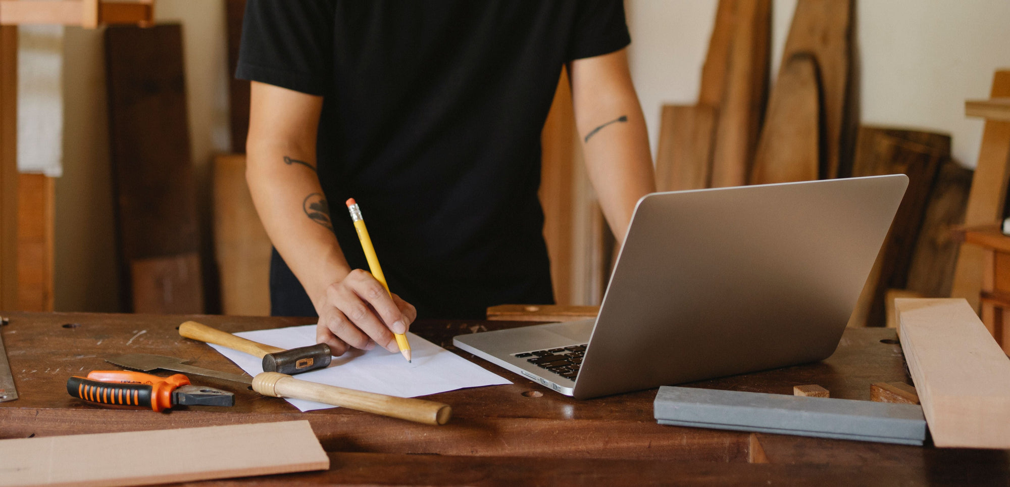 A person in a black t-shirt stands at a wooden workbench, writing with a yellow pencil on white paper next to an open laptop.