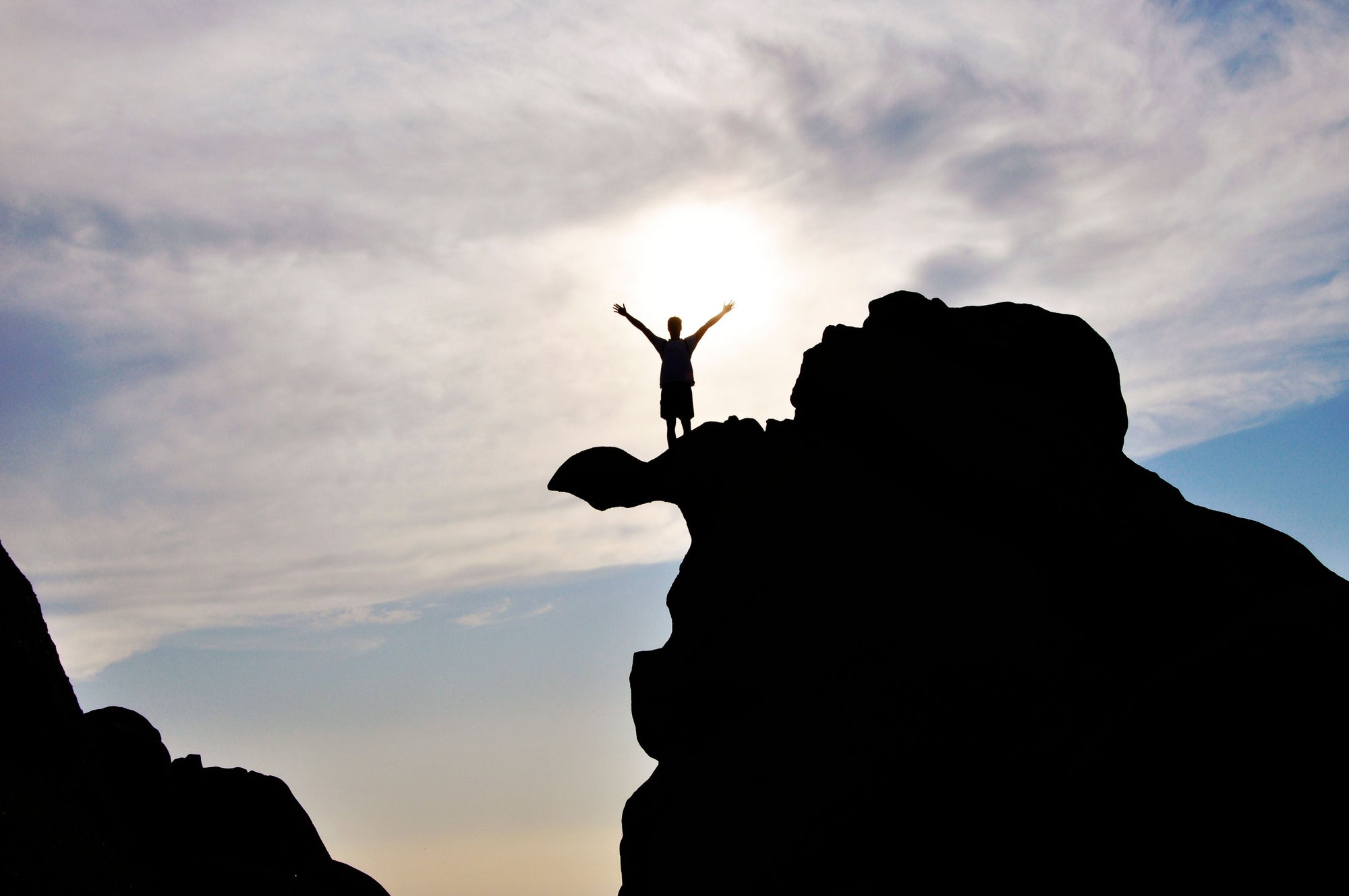 A silhouetted person stands triumphantly with arms raised atop a rugged rock formation, backlit by the sun against a cloudy sky.