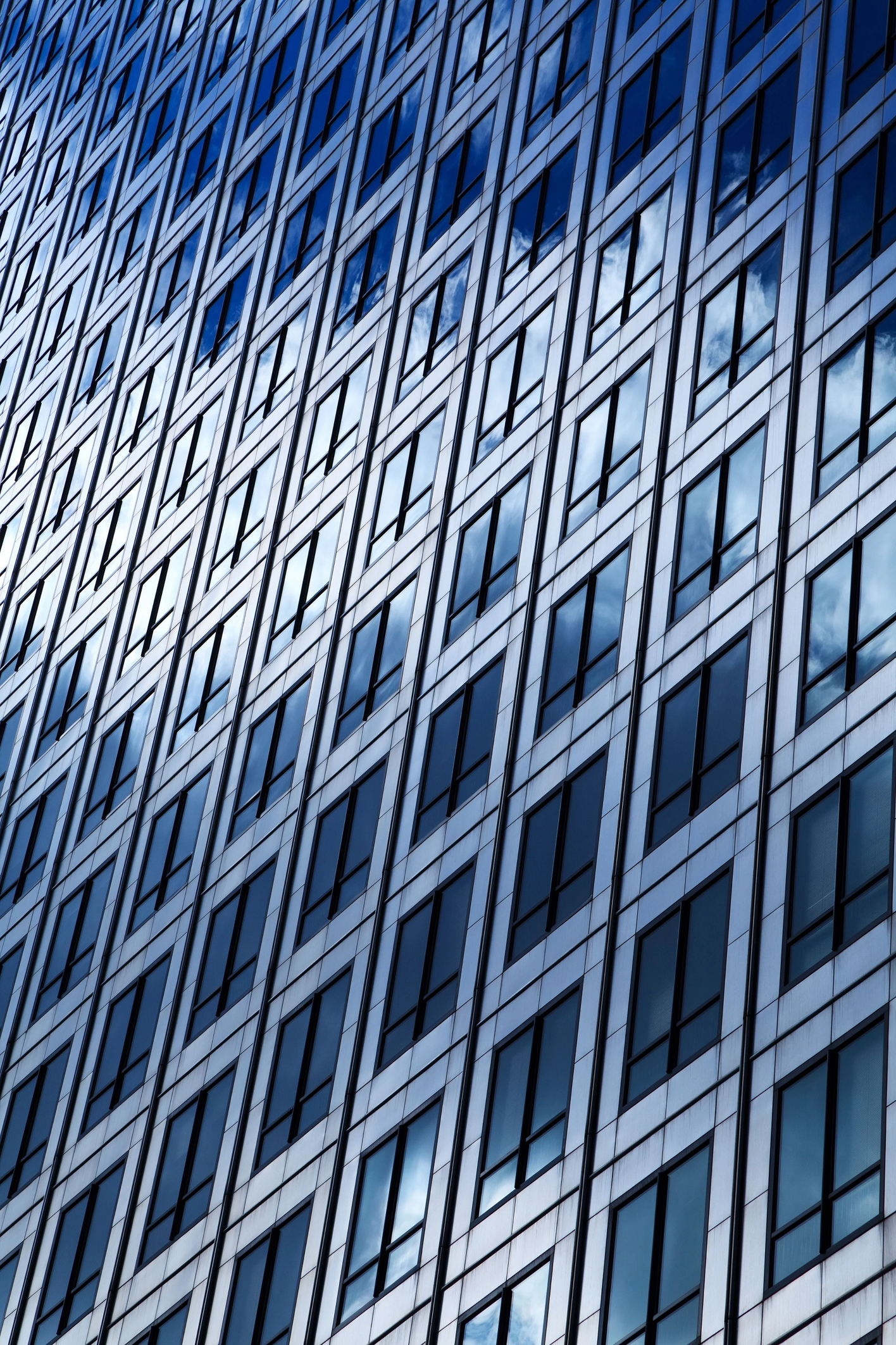 Close-up view of a high-rise office building's glass and steel facade, with rows of windows reflecting a blue sky and passing clouds in a repeating grid pattern.