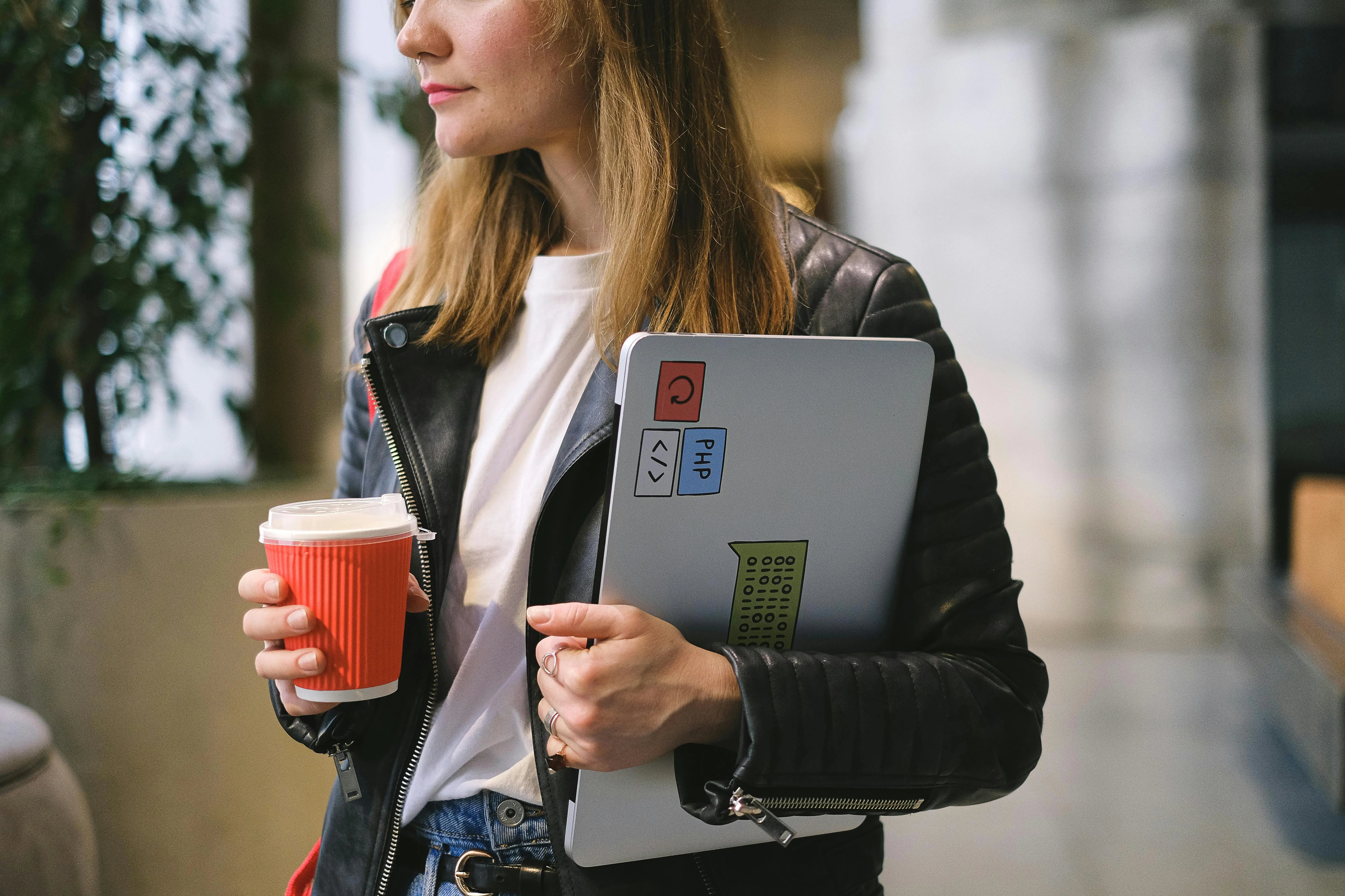 A casually dressed woman holding a laptop with tech-related stickers, including PHP and code symbols, in one hand and a red takeaway coffee cup in the other
