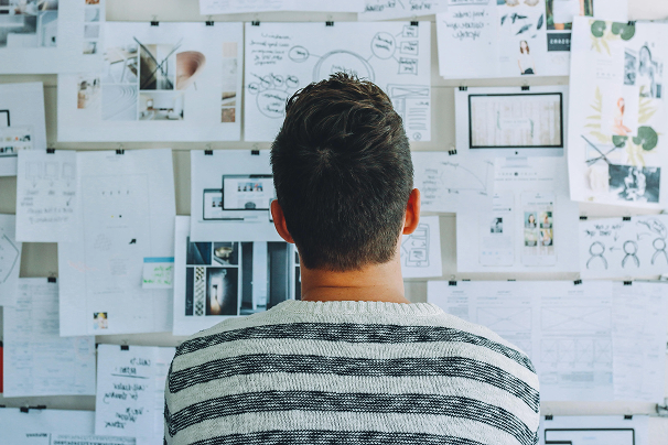 A man with short hair viewed from behind, standing in front of a wall covered with sketches, notes, and design concepts.