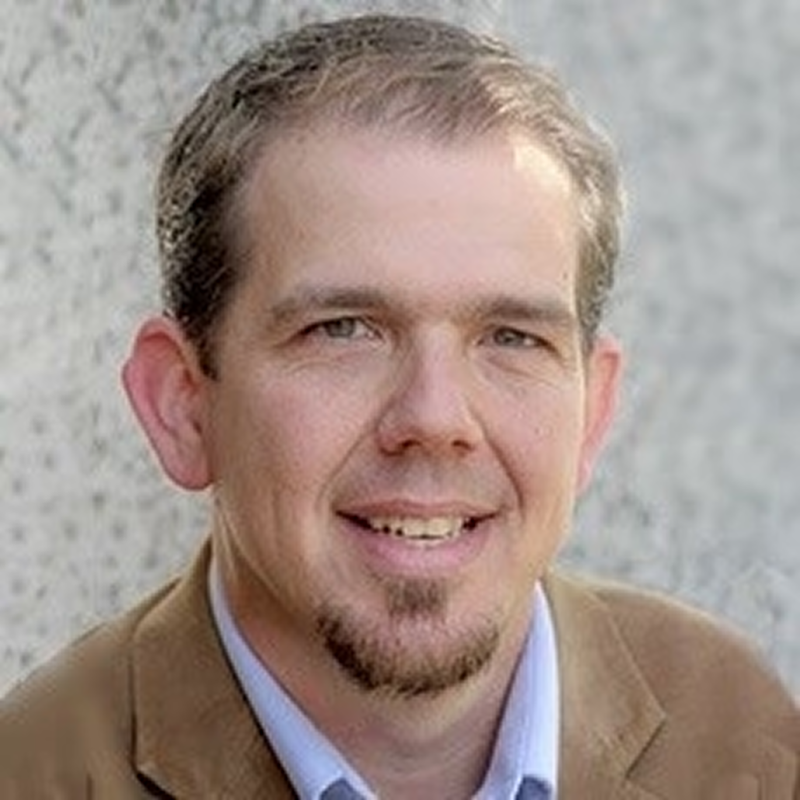 Ashby smiling gently, with short hair and a goatee, wearing a tan blazer and light blue shirt.