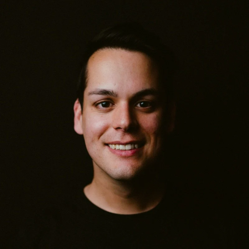 Behzod smiling against a dark background, with short dark hair and a black shirt.