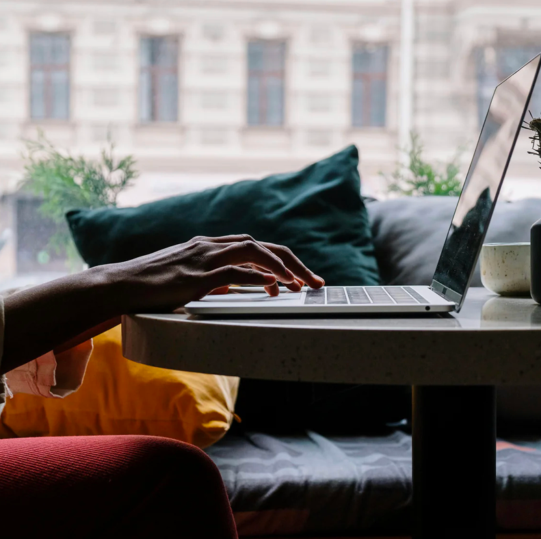 A person typing on a laptop at a small round table in a cozy window-side seating area with green and yellow cushions, plants, and a coffee cup nearby.