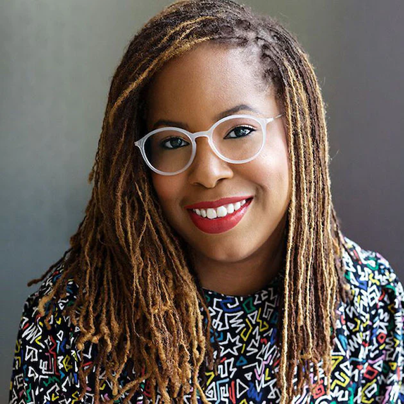 Patrice smiling at the camera, with long locs, white-framed glasses, and a colorful patterned top.