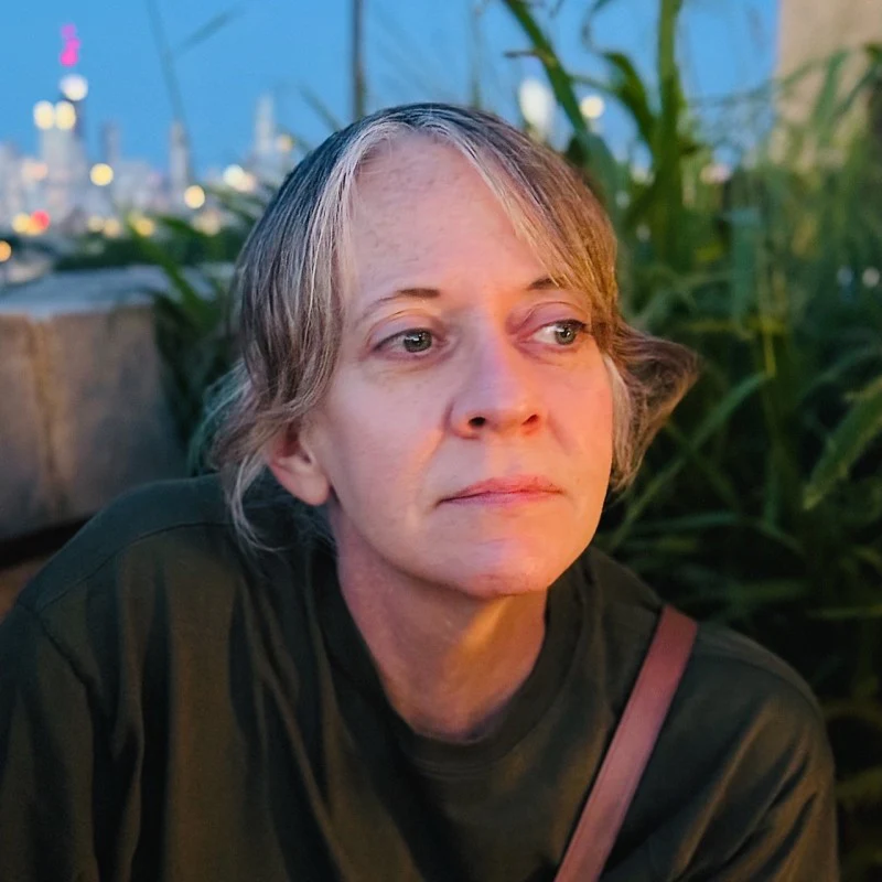 Rachael looking off to the side at dusk, with shoulder-length blonde hair and an olive shirt, a city skyline behind