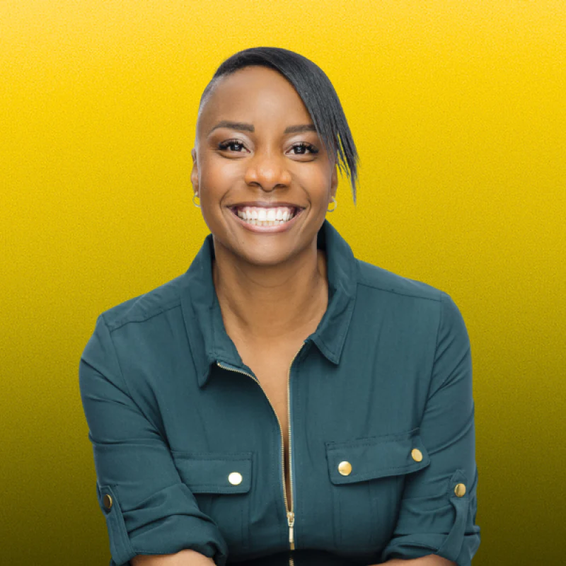 Vivianne smiling warmly against a bright yellow background, with a short dark style and a teal zip-up top.