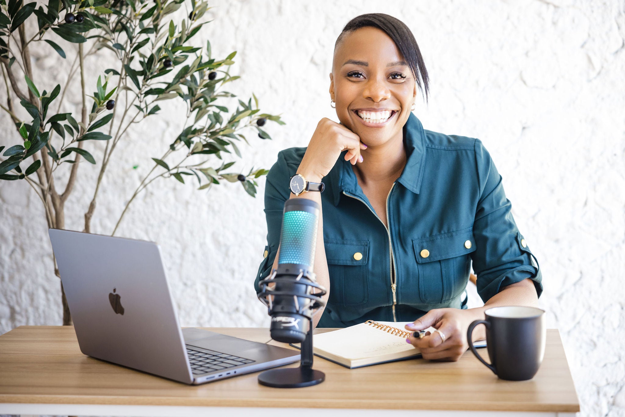 Vivianne smiling in a teal button-up dress resting her chin on her hand, seated at a wooden desk with a laptop, a podcast microphone, an open spiral notebook, and a dark mug, with a leafy olive tree and white textured wall behind her.