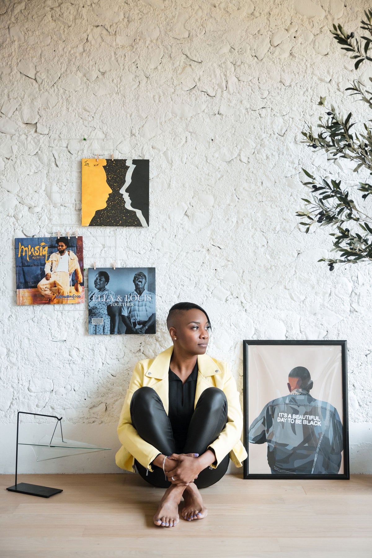 Vivianne with a shaved-side haircut sits barefoot on a light wood floor against a white textured stone wall, wearing a pale yellow leather jacket over black clothing. She's surrounded by framed art and album covers, including Ella Fitzgerald and Louis Armstrong's "Ella & Louis Together," a Musiq Soulchild album, and a large framed print of a figure in a denim jacket reading "It's a Beautiful Day to Be Black." A small olive branch enters the frame at top right.